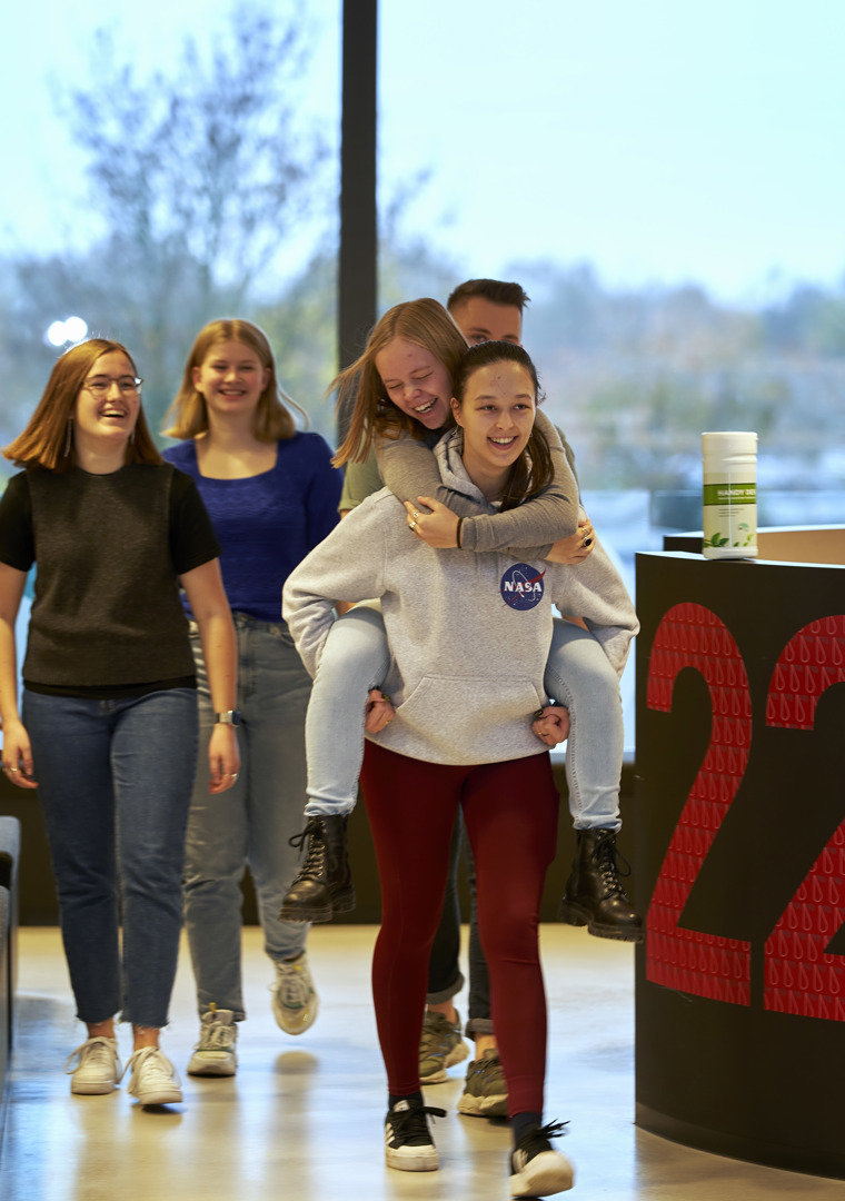Fem skoleelever kommer gående i et fællesområde. Den forreste elev bærer en anden elev på ryggen - de griner og ser glade ud.