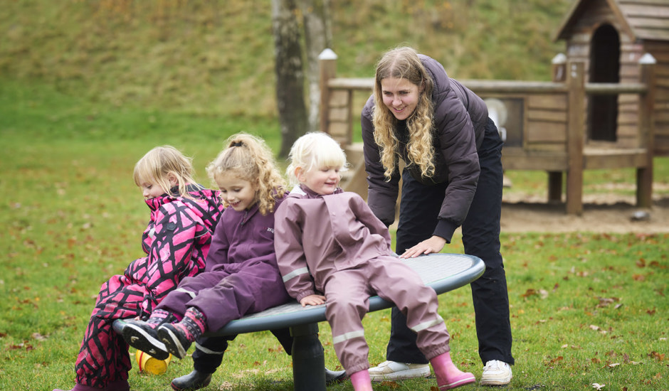 En pædagogisk assistent hjælper tre børn, som sidder tæt sammen på et vippebræt på legepladsen. Alle smiler og er iført regnsæt.